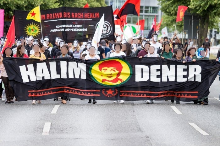 Eine Demonstration auf der Straße. Vorne ist ein großes Transparent mit der Aufschrift "Halim Dener" und dem Gesicht von Halim Dener. Im Hintergrund ist ein Transparent mit der Aufschrift "Von Rojava bis nach Berlin - Kommunales Leben aufbauen und verteidigen". Einige Menschen tragen das Gesicht von Halim Dener als Maske.